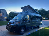 This RV rental is a dark gray camper van with a raised pop-top, parked on a driveway with blue ground lights at twilight.