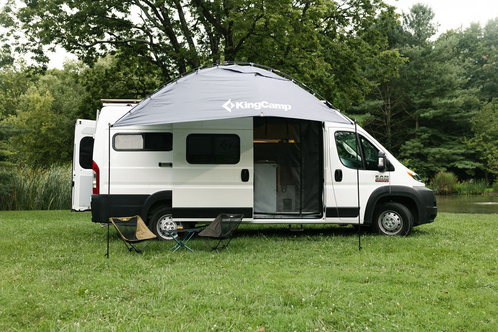 White RV rental with a KingCamp awning extended, chairs, and table on grass by woods and water.