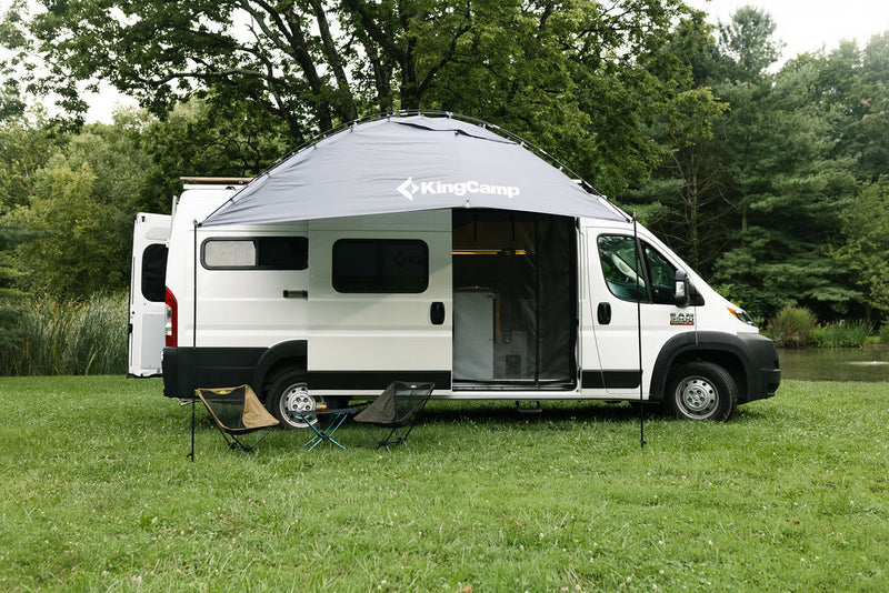 White RV rental with a KingCamp awning extended, chairs, and table on grass by woods and water. — photo 1