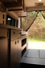 Modern RV rental interior: compact kitchen, storage, and open back door revealing a peaceful wooded outdoor scene.