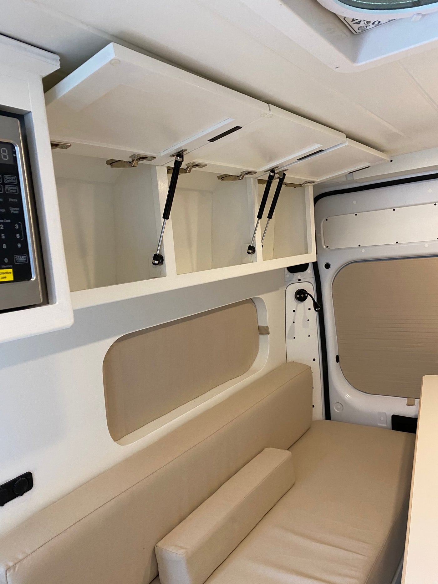 Inside an RV rental van: bright white overhead cabinets with open doors, microwave, & neutral cushioned bench seating. — photo 10