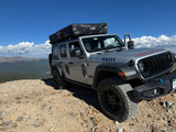 A silver Jeep Wrangler 4-door, an off-road rv rental, with a rooftop tent on a rocky mountain peak. Stunning mountain views.