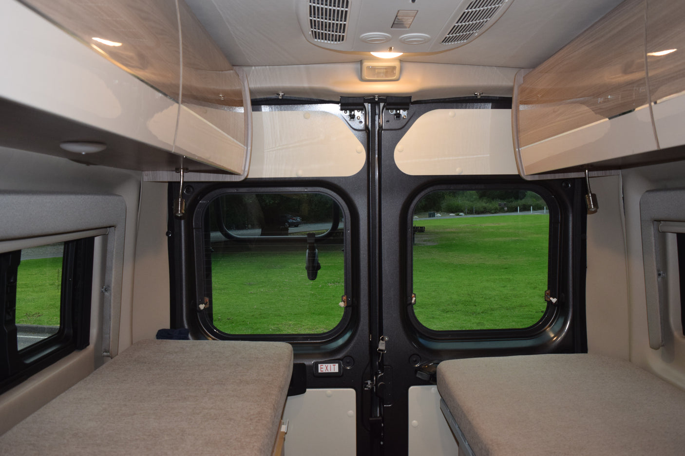 RV rental interior featuring cozy beds, overhead storage, and a scenic view through the rear doors to a grassy field.