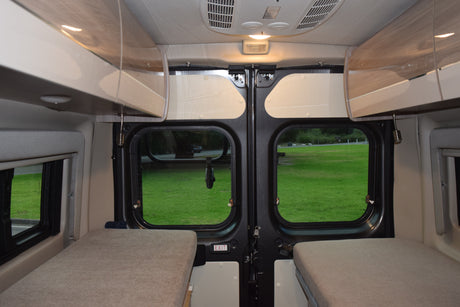 RV rental interior featuring cozy beds, overhead storage, and a scenic view through the rear doors to a grassy field.