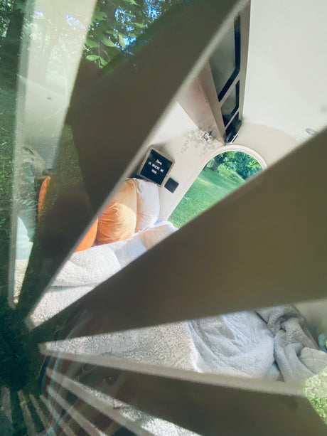Experience a cozy RV rental: complete with bed, pillows, and an arched window looking out to nature. "Home is where we park."