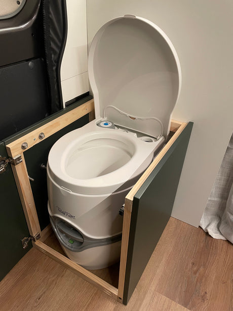 White portable toilet, lid open, integrated into a custom cabinet within an RV rental. Essential for comfortable van life.