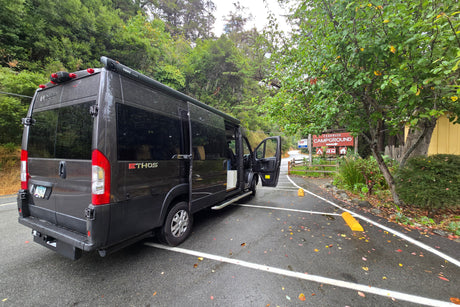 Dark gray rv rental camper van with open door parked at Fernwood Campground entrance, surrounded by lush green trees.