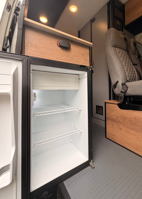 Open, empty refrigerator in a modern RV rental camper van, showing wire shelves, wood cabinetry, and a passenger seat.