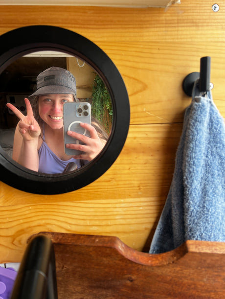 Smiling in an **rv rental**, a woman in a hat takes a mirror selfie with a peace sign. Cozy wooden interior with blue towel.