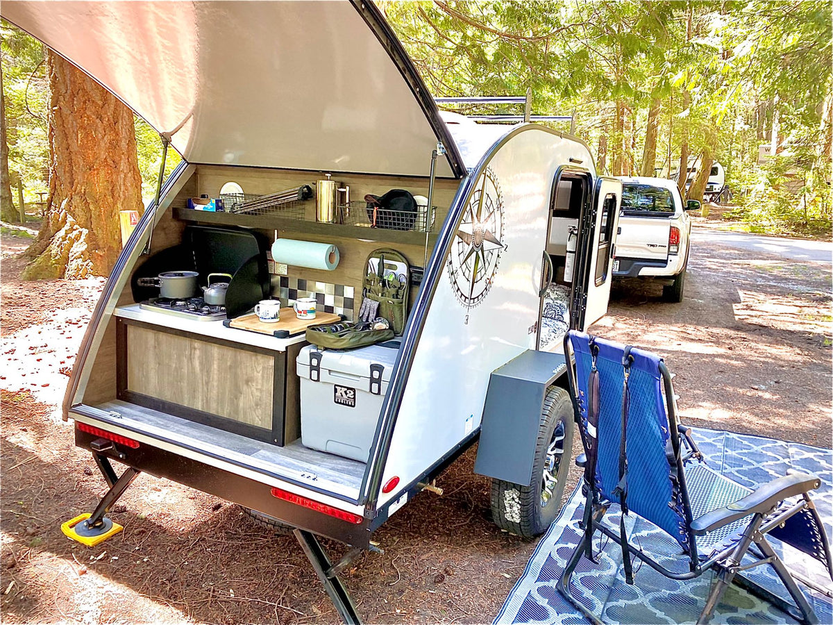 A modern teardrop RV rental setup with a fully equipped outdoor kitchen, awning, and camping chairs in a forest campground. — photo 3