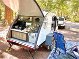 A modern teardrop RV rental setup with a fully equipped outdoor kitchen, awning, and camping chairs in a forest campground.