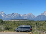 A gray RV rental on a road, with stunning mountains & green landscape under a clear blue sky. Ideal for road trips.