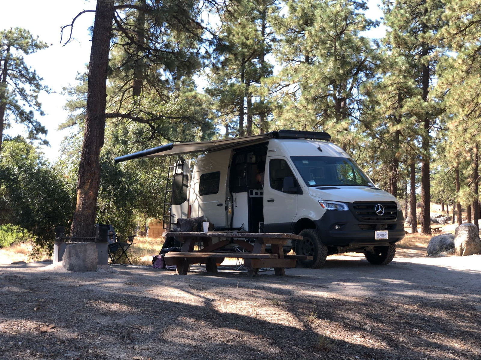 A white Mercedes-Benz Sprinter RV rental parked at a serene campsite with an extended awning among tall pines.
