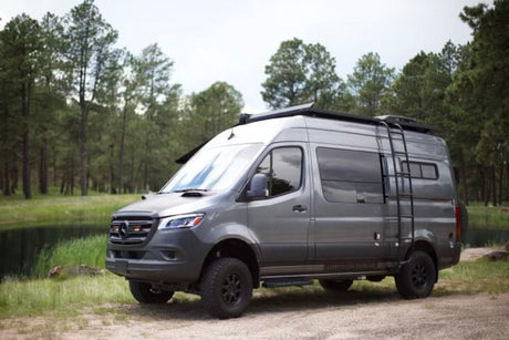 Rugged grey Mercedes-Benz Sprinter RV rental with off-road tires & roof ladder, by a forest pond.