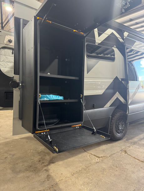 Exterior storage locker open on a custom adventure van, great for RV rental gear, with shelves & fold-down table.