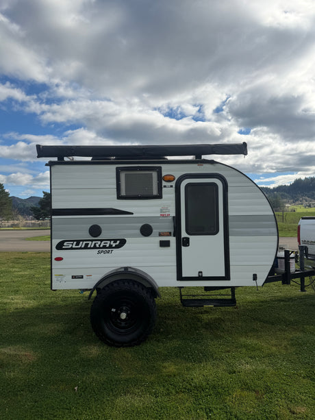 A compact Sunray Sport travel trailer, ideal for an rv rental, sits on grass with mountains & lake in background under a cloudy sky.