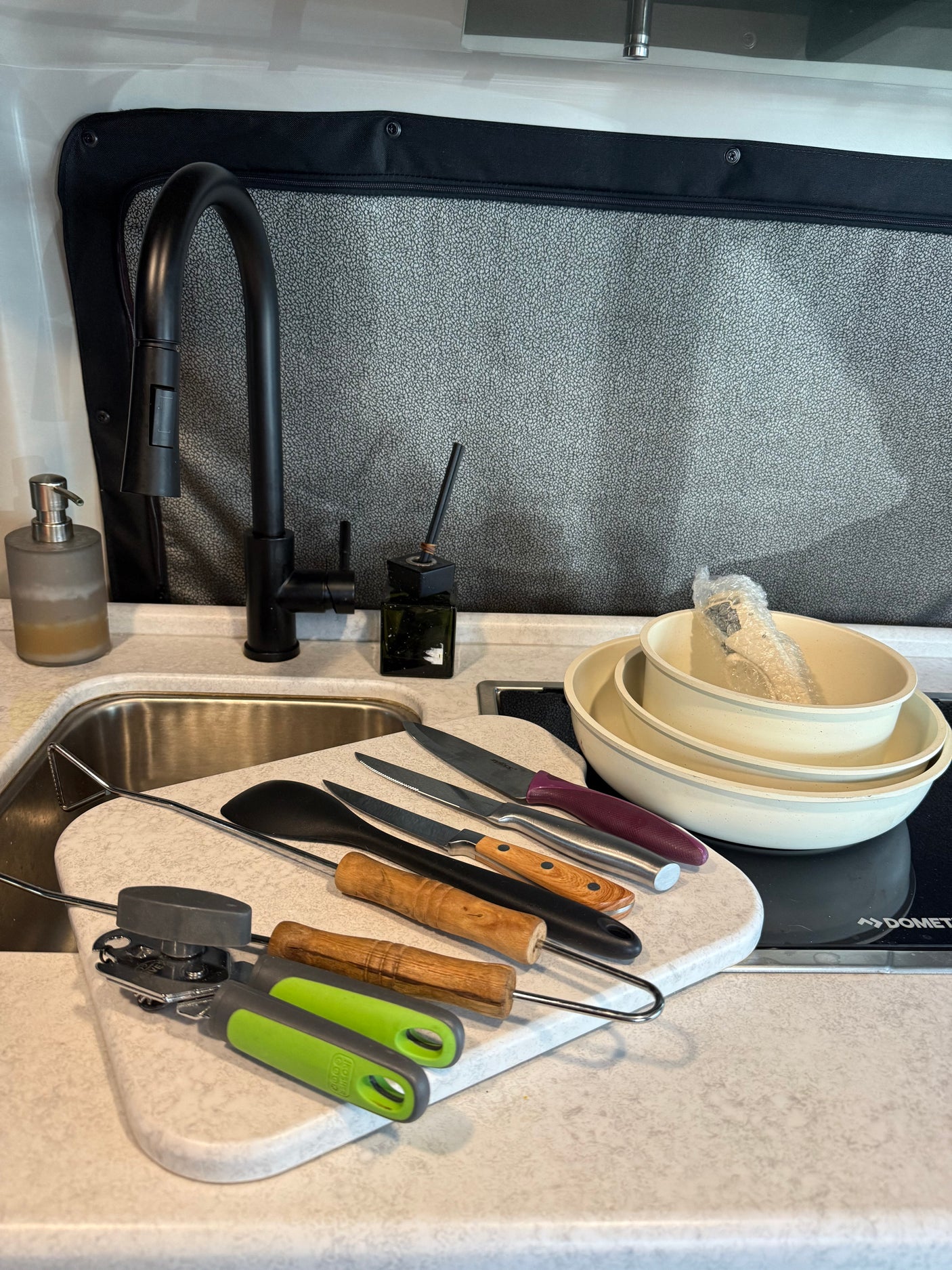 RV rental kitchen: black faucet, sink, cutting board with knives, spatula, can opener, tongs, bowls, and cooktop.