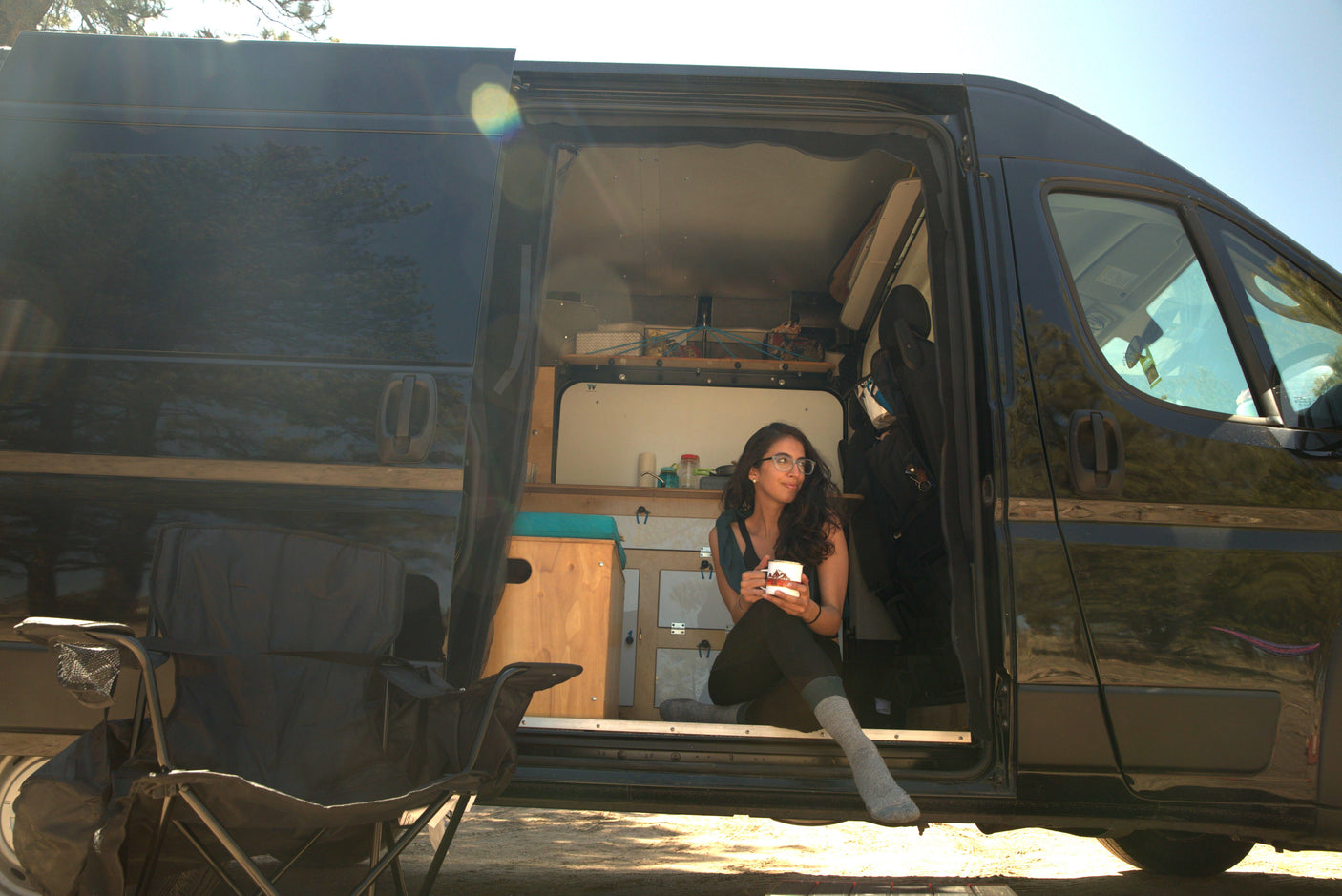 Happy woman in glasses enjoying coffee inside an open black RV rental camper van. Perfect for adventure & van life.