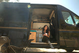 Happy woman in glasses enjoying coffee inside an open black RV rental camper van. Perfect for adventure & van life.