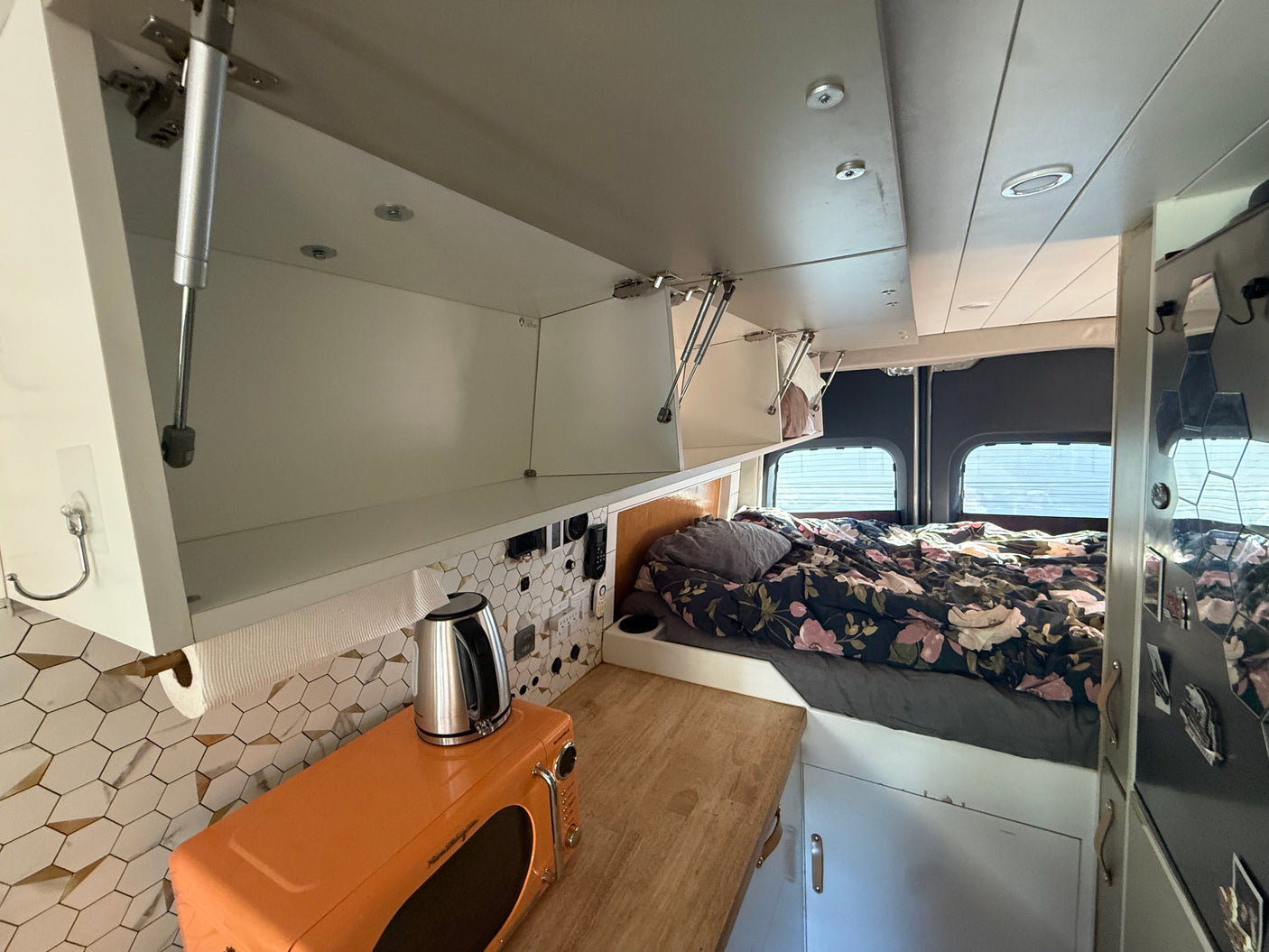 Inside an rv rental: compact camper van kitchen and bed area. White open overhead cabinets, orange microwave, kettle, and floral bedding.