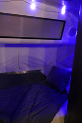 Inside an RV rental: a dimly lit bedroom with blue ambient lights, a made bed, pillows, and a window. Ready for your trip.