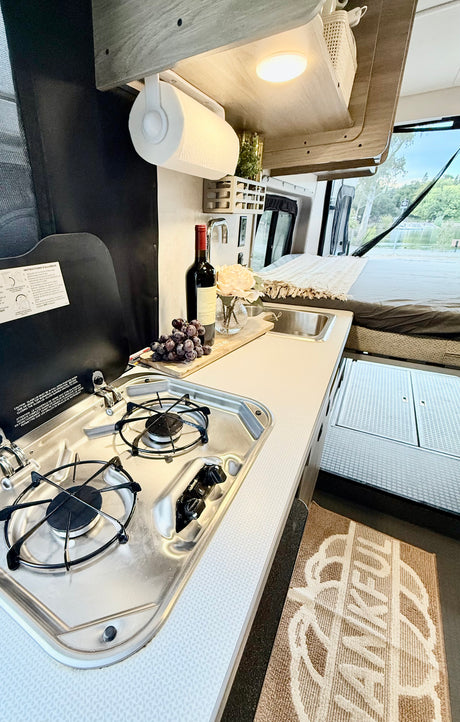 Cozy RV rental kitchen: stove, sink, paper towels, wine, and bed with a scenic lake view. Enjoy compact living.