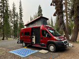 This vibrant red Winnebago Solis rv rental is parked at a forest campsite, pop-top extended, with an open door.