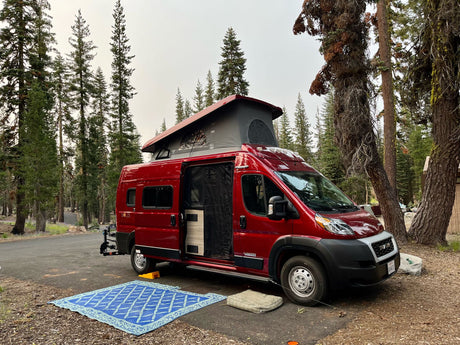 This vibrant red Winnebago Solis rv rental is parked at a forest campsite, pop-top extended, with an open door.