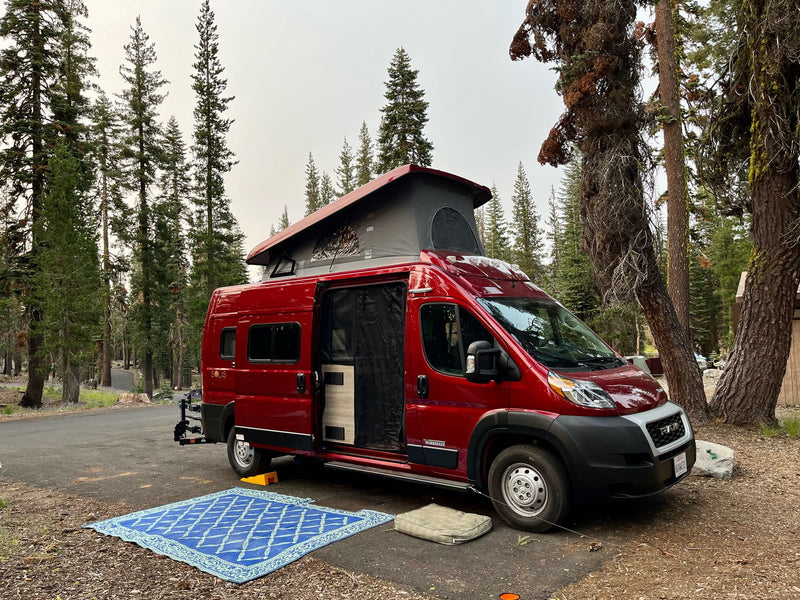  surrounded by pines under a blue sky.|This vibrant red Winnebago Solis rv rental is parked at a forest campsite — photo 3