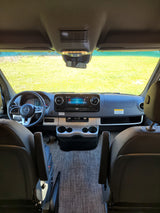 Inside a modern RV rental: a detailed view of the dashboard, Mercedes-Benz steering wheel, and infotainment screen, facing vibrant green fields.