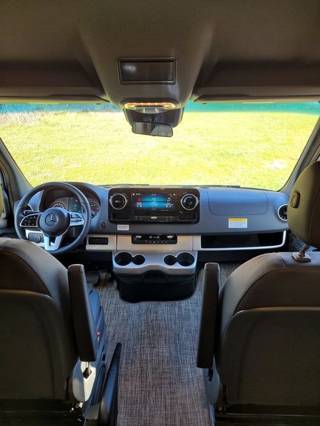 Inside a modern RV rental: a detailed view of the dashboard, Mercedes-Benz steering wheel, and infotainment screen, facing vibrant green fields.