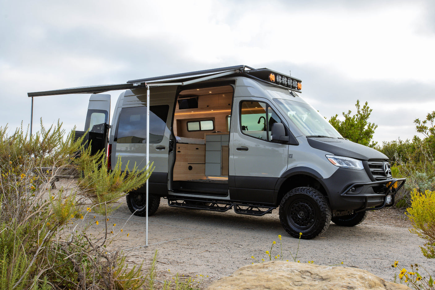 Ready for adventure! Custom light grey Mercedes Sprinter rv rental with off-road tires, extended awning, and open, cozy interior.