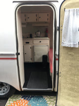 Open door view of a compact RV rental interior, showing a kitchen with sink/stove, cabinets, and a seating area.