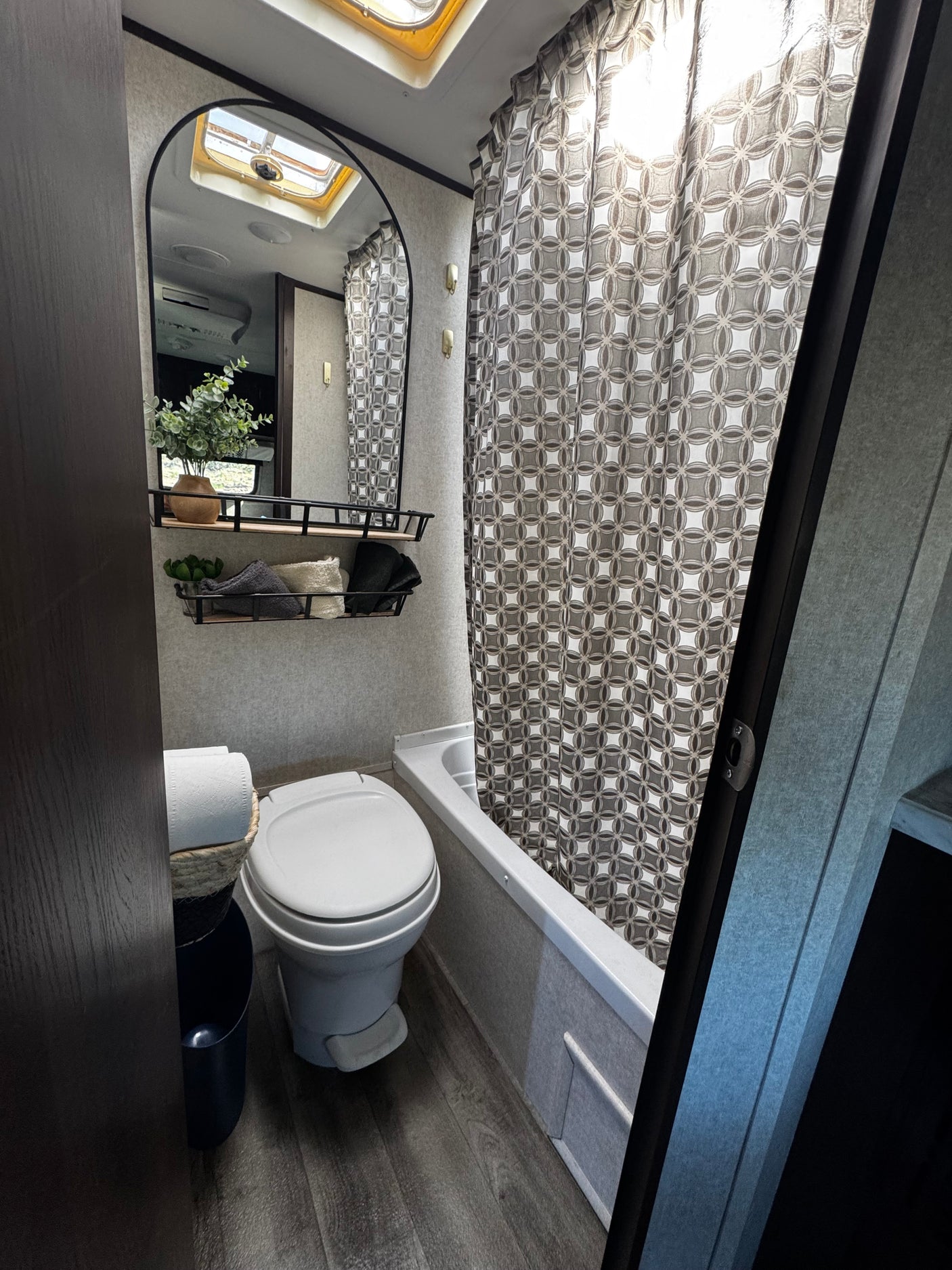 Clean RV rental bathroom featuring toilet, shower/tub, mirror, skylight, and decorative storage. Ideal for travel.