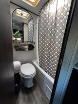 Clean RV rental bathroom featuring toilet, shower/tub, mirror, skylight, and decorative storage. Ideal for travel.