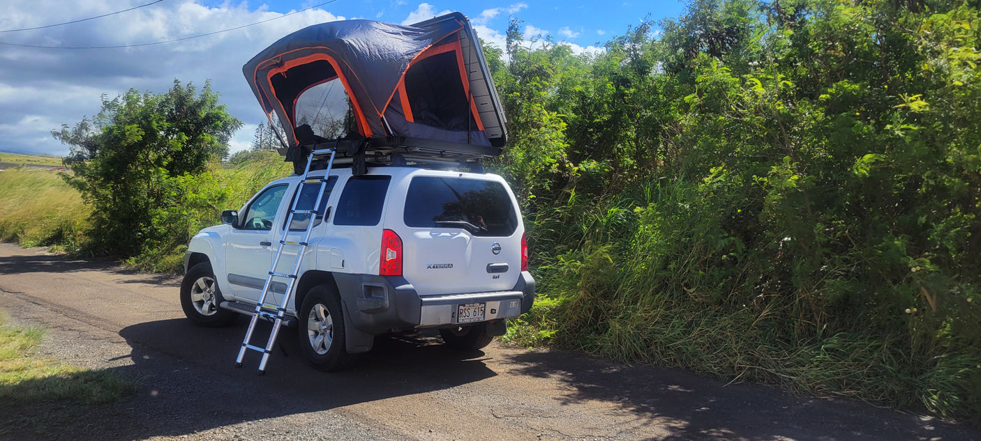 White Nissan Xterra SUV with a deployed roof-top tent & ladder, parked on a road. Great RV rental for camping or adventure travel.
