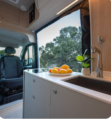 This modern RV rental kitchen has a sleek counter, sink, fruit bowl, and a stunning view of trees through a large window.