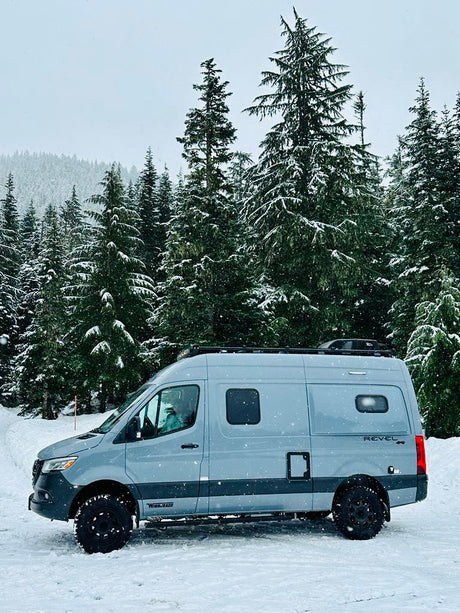 Gray Winnebago Revel camper van, perfect for rv rental, parked in a snowy forest amidst tall evergreens.