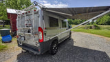 Parked silver RV rental camper van with a large awning extended, on a gravel drive with green fields.