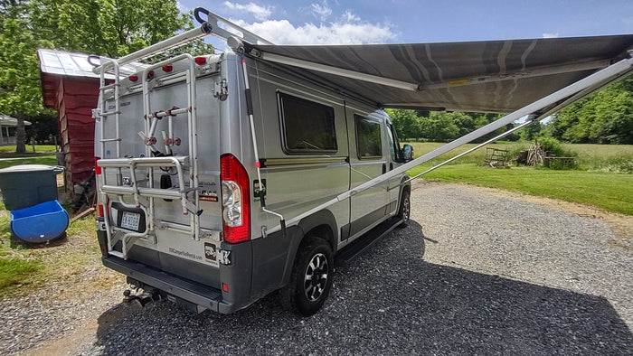 Parked silver RV rental camper van with a large awning extended, on a gravel drive with green fields.