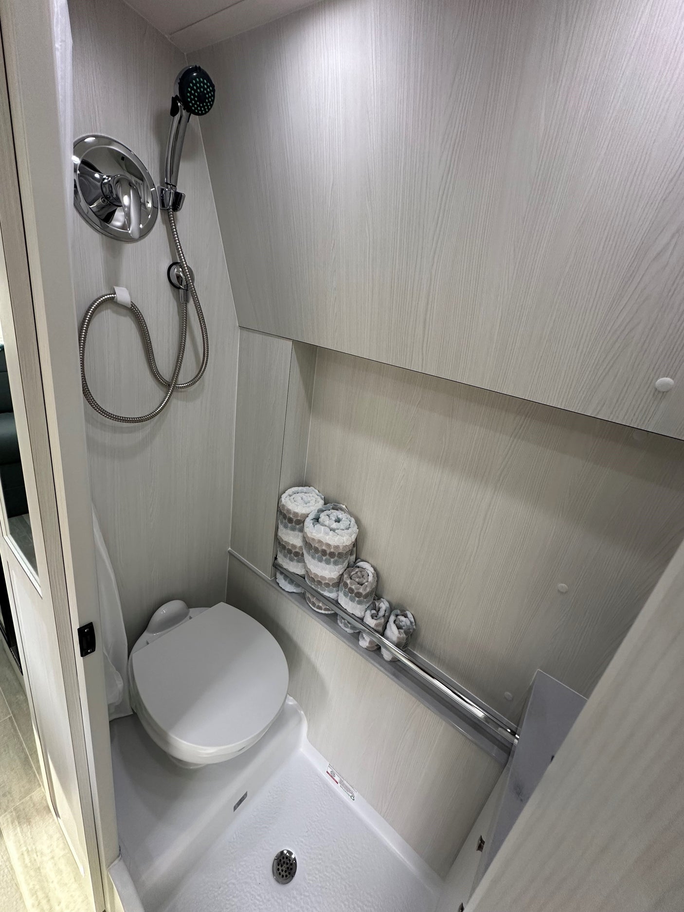 RV rental's compact bathroom: shower, toilet, and rolled towels on a shelf. Light-toned, efficient design.