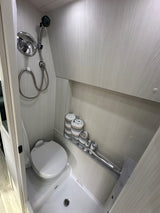RV rental's compact bathroom: shower, toilet, and rolled towels on a shelf. Light-toned, efficient design.