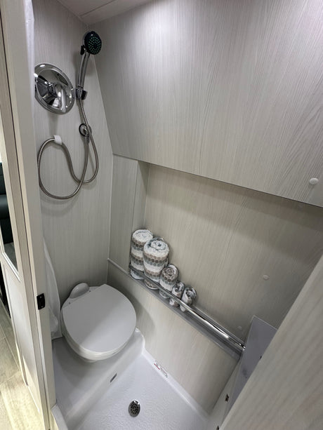 RV rental's compact bathroom: shower, toilet, and rolled towels on a shelf. Light-toned, efficient design.