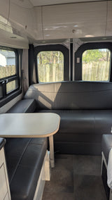 An rv rental's dinette area features black seating, a light table, overhead storage, and exterior views through windows.