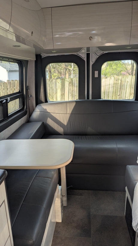 An rv rental's dinette area features black seating, a light table, overhead storage, and exterior views through windows.
