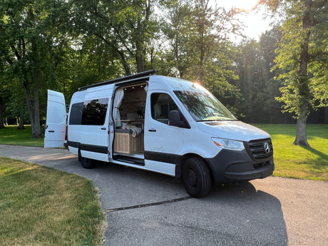 White Mercedes Sprinter van RV rental with open doors showcasing its custom interior, parked outdoors amidst green trees.