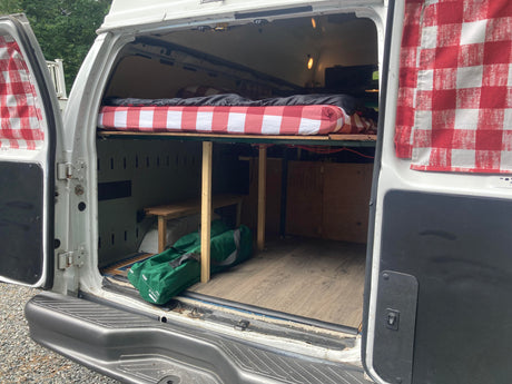 Back view of a DIY camper van conversion with a raised bed, storage, and checkered curtains, ideal for RV rental travel.