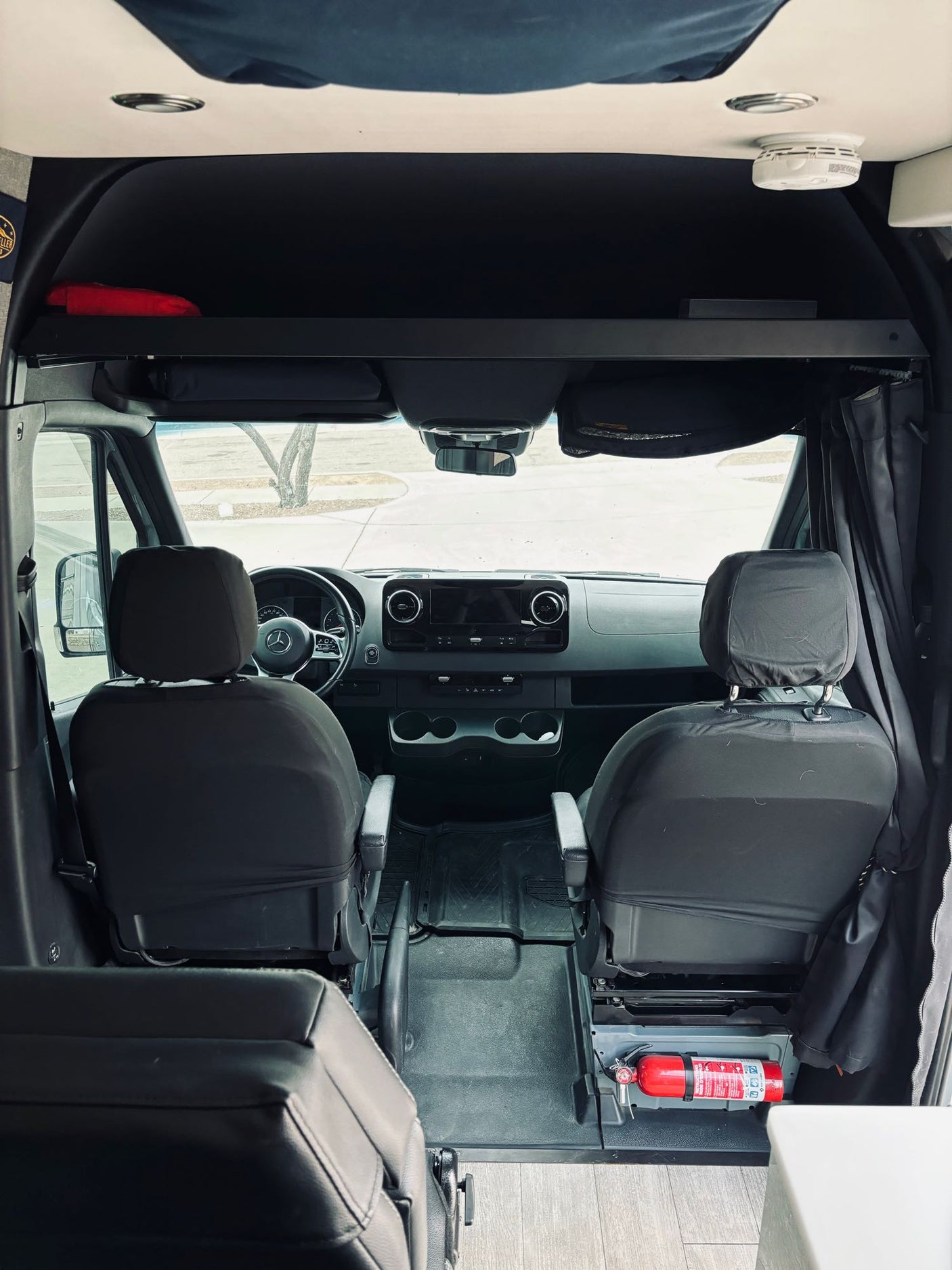 View of an RV rental's front cab from the rear, showing seats, dashboard, overhead shelf, and fire extinguisher.