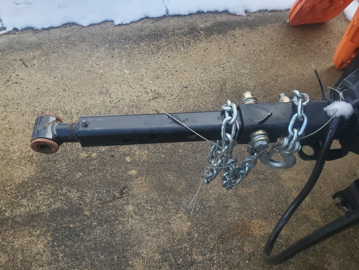 RV rental trailer hitch with safety chains on concrete pavement, featuring attachment bolts and a safety cable. Snow is present.