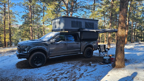 Grey Ford Raptor with an extended rooftop tent, an ideal RV rental alternative for snowy forest camping adventures.